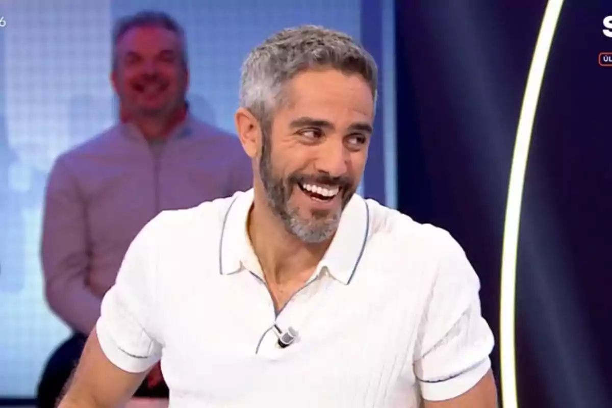 Roberto Leal de cabello corto y barba canosa sonríe mientras participa en un programa de televisión con fondo azul y otra persona desenfocada detrás. Roberto Leal de cabello corto y barba canosa sonríe mientras participa en un programa de televisión con fondo azul y otra persona desenfocada detrás.