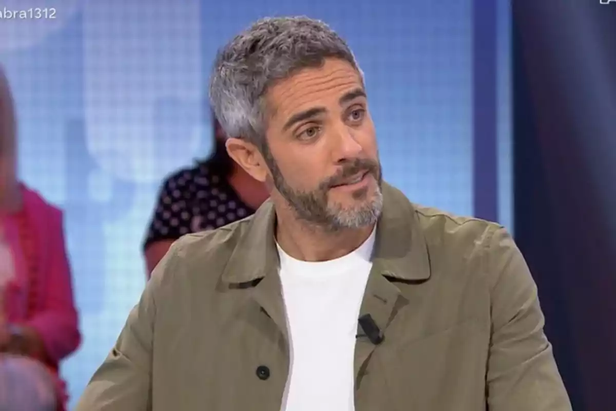 Home de cabell curt i barba canosa vestit amb jaqueta beix i samarreta blanca participant en un programa de televisió amb fons desenfocat i persones assegudes al darrere Home de cabell curt i barba canosa vestit amb jaqueta beix i samarreta blanca participant en un programa de televisió amb fons desenfocat i persones assegudes al darrere