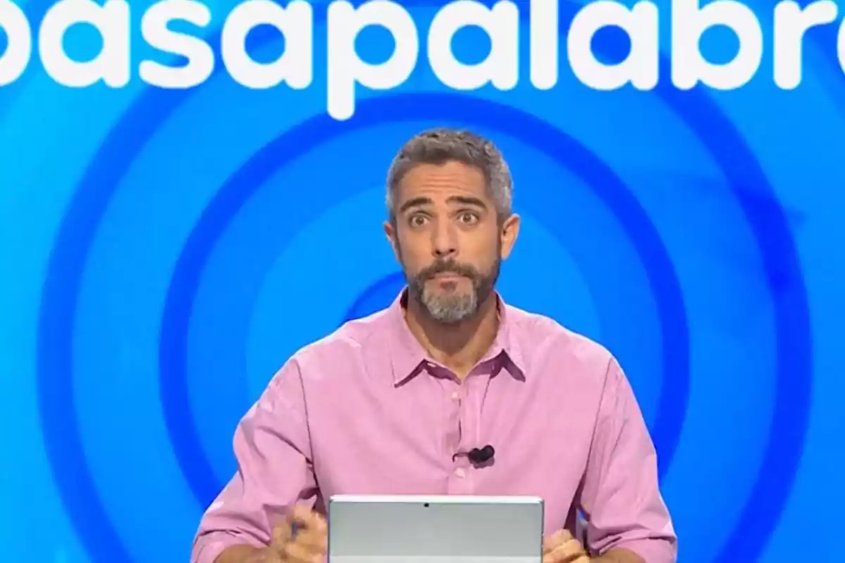 Home amb barba i camisa rosa assegut davant d’una pantalla blava amb el logotip d’un programa de televisió de concursos. Home amb barba i camisa rosa assegut davant d’una pantalla blava amb el logotip d’un programa de televisió de concursos.