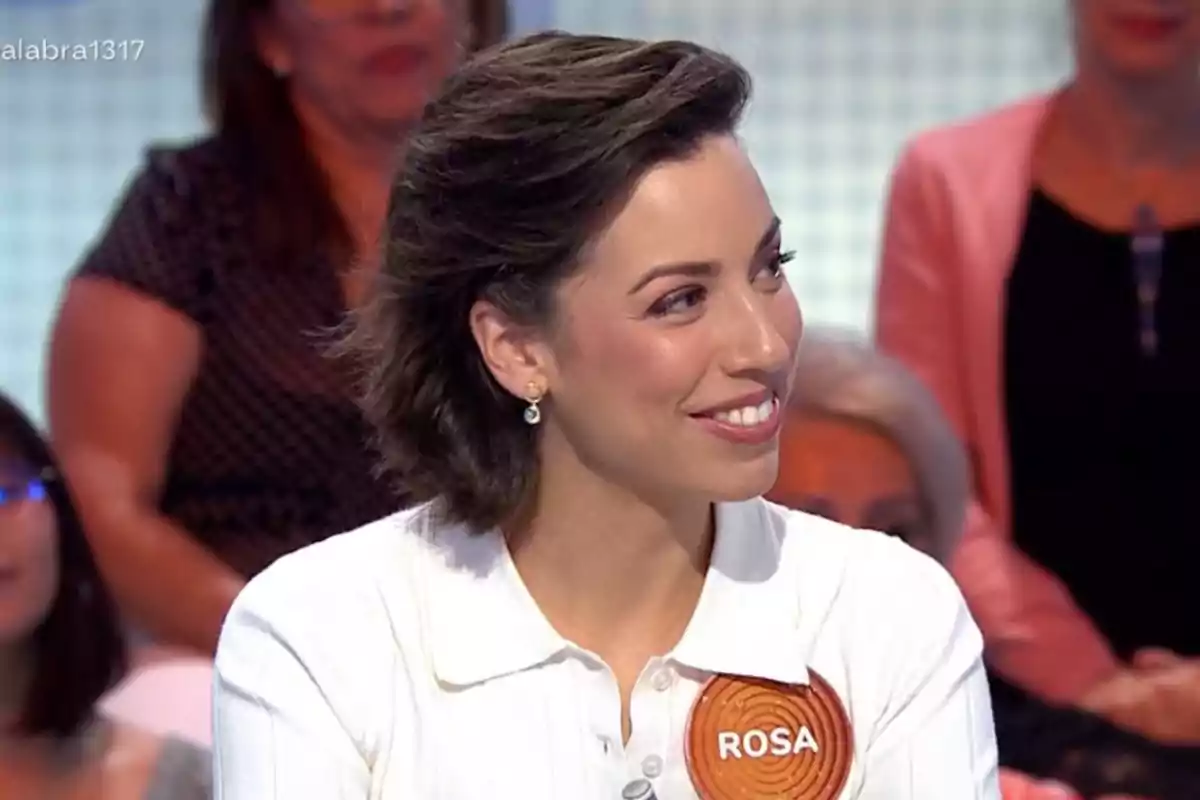 Dona de cabell curt i fosc somriu mentre participa en un programa de televisió, porta una camisa blanca i una placa amb el nom Rosa. Dona de cabell curt i fosc somriu mentre participa en un programa de televisió, porta una camisa blanca i una placa amb el nom Rosa.