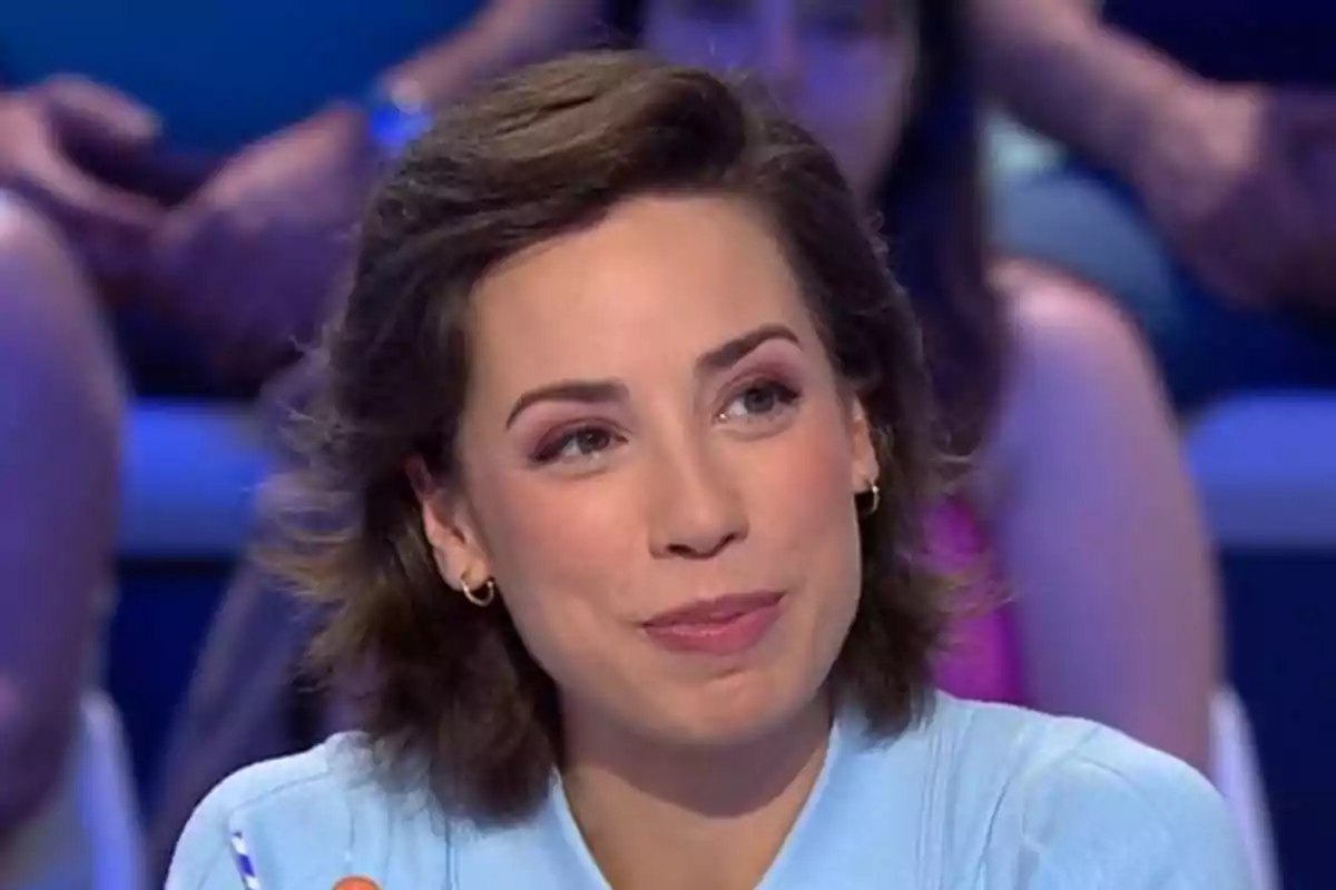 Rosa de cabello corto y castaño con expresión sonriente, usando una camisa azul claro y pendientes dorados, sentada en un estudio de televisión con personas desenfocadas al fondo
