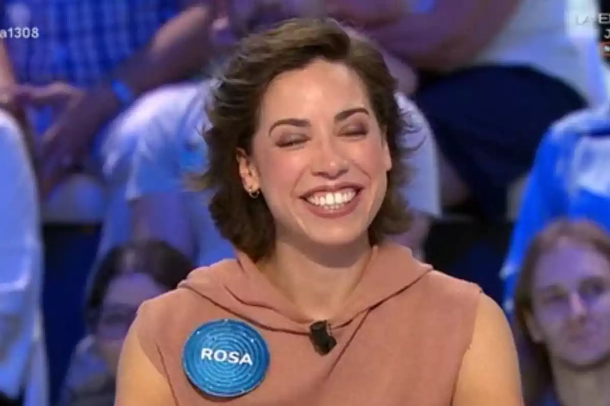 Rosa sonriente con cabello corto y castaño, lleva un micrófono y una chapa azul con el nombre Rosa, está en un programa de televisión con público de fondo. Rosa sonriente con cabello corto y castaño, lleva un micrófono y una chapa azul con el nombre Rosa, está en un programa de televisión con público de fondo.