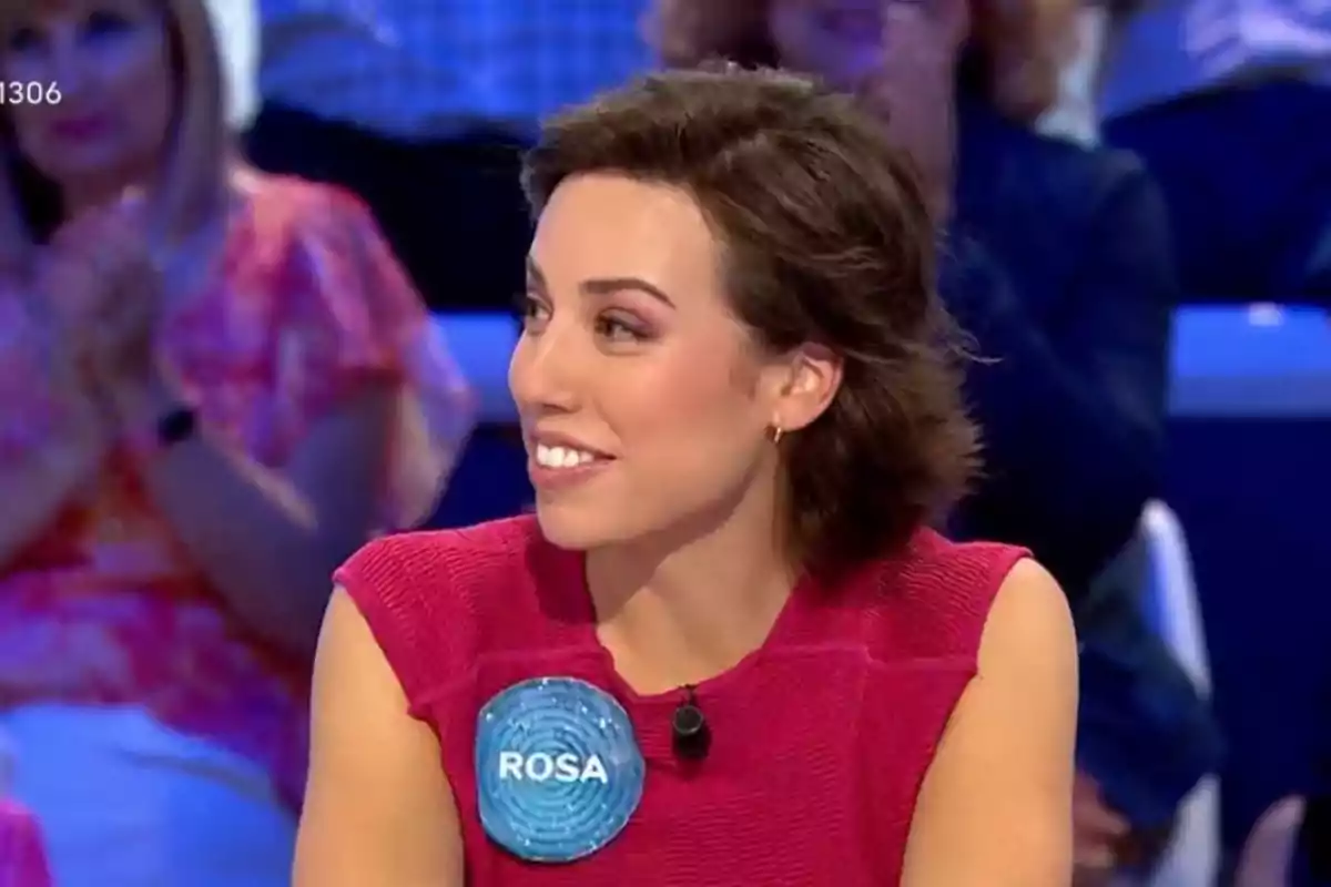 Rosa joven con cabello corto y oscuro, vestida con una blusa rosa y una placa azul que dice Rosa, sentada en un plató de televisión con personas desenfocadas al fondo. Rosa joven con cabello corto y oscuro, vestida con una blusa rosa y una placa azul que dice Rosa, sentada en un plató de televisión con personas desenfocadas al fondo.