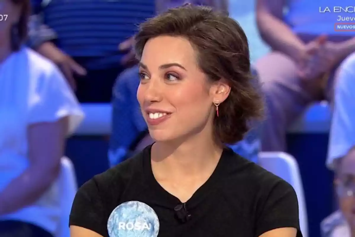 Dona jove de cabell curt i fosc somriu mentre participa en un programa de televisió, porta una samarreta negra i una acreditació blava amb el nom Rosa. Dona jove de cabell curt i fosc somriu mentre participa en un programa de televisió, porta una samarreta negra i una acreditació blava amb el nom Rosa.