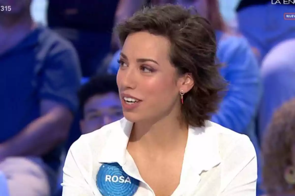 Rosa de cabello corto y castaño con camisa blanca y un pin azul con el nombre Rosa participando en un programa de televisión con público de fondo Rosa de cabello corto y castaño con camisa blanca y un pin azul con el nombre Rosa participando en un programa de televisión con público de fondo