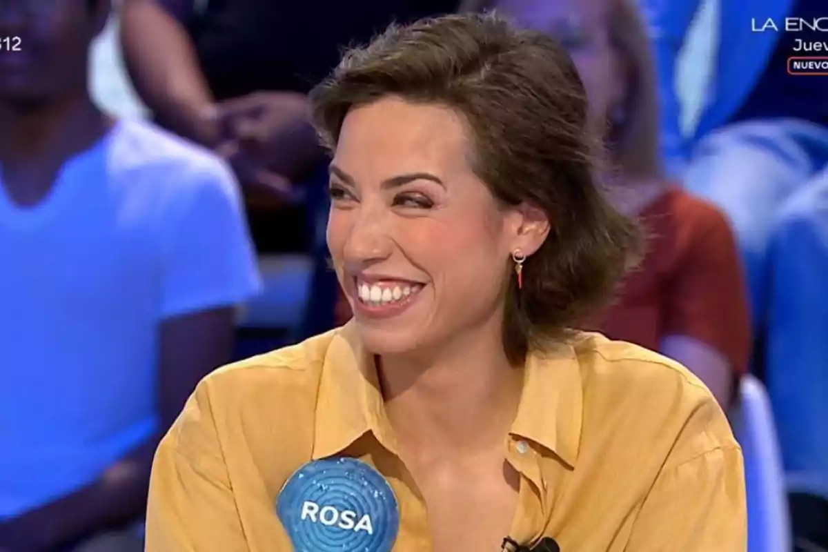 Dona somrient amb camisa groga participant en un programa de televisió amb una xapa blava que diu ROSA Dona somrient amb camisa groga participant en un programa de televisió amb una xapa blava que diu ROSA