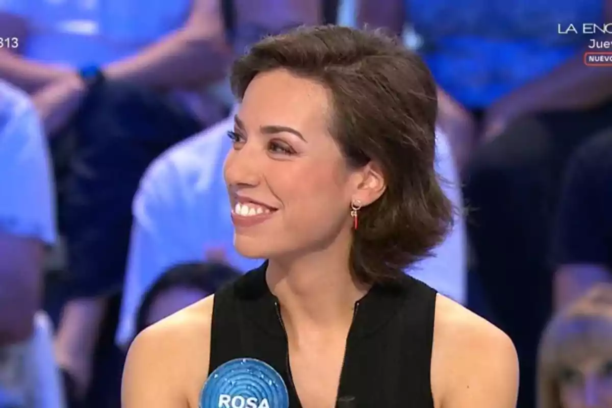Rosa joven de cabello corto y oscuro sonríe mientras participa en un programa de televisión, lleva un vestido negro y un pendiente rojo Rosa joven de cabello corto y oscuro sonríe mientras participa en un programa de televisión, lleva un vestido negro y un pendiente rojo