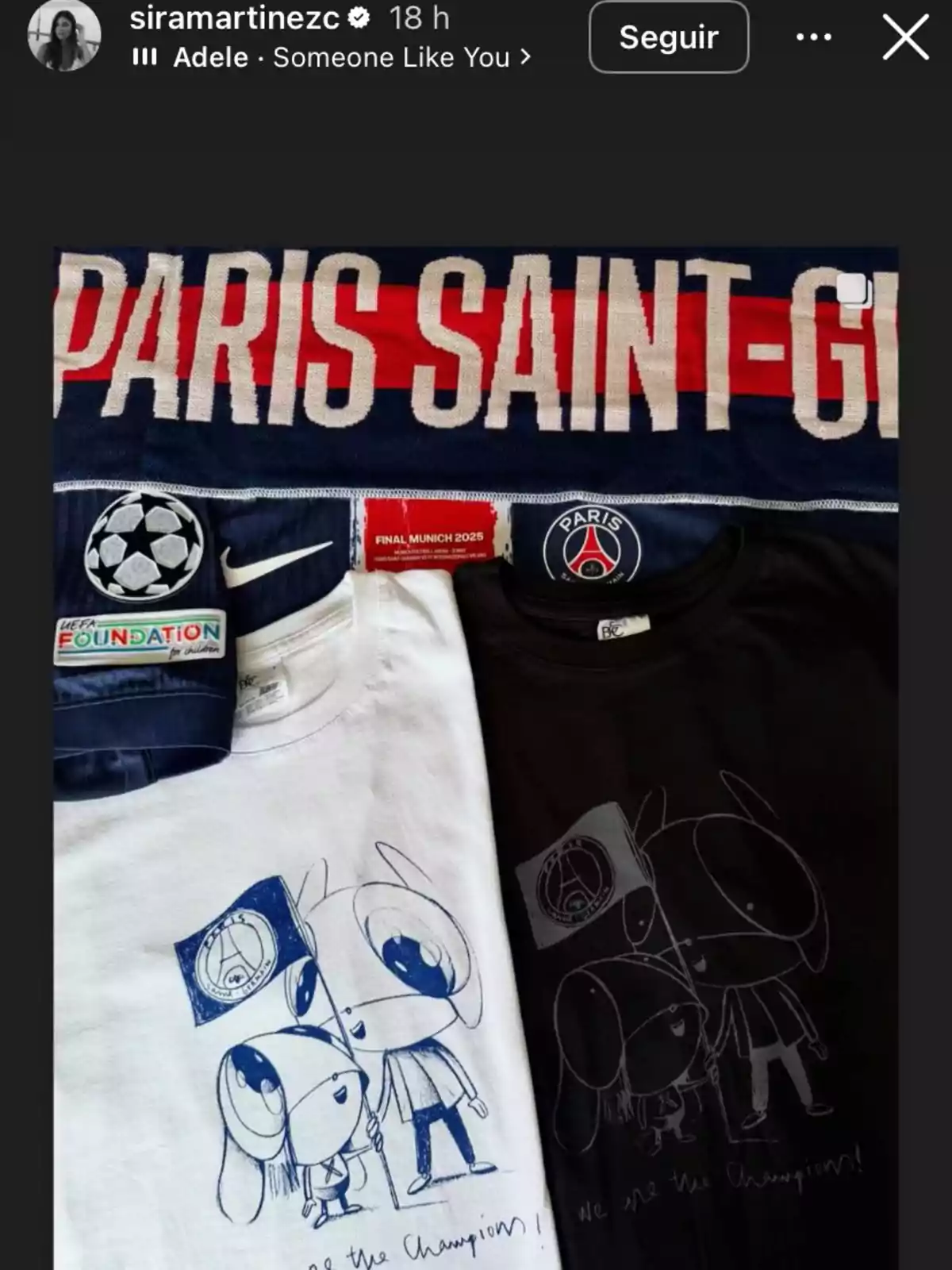 Dos camisetas, una blanca y una negra, con un dibujo de dos personajes sosteniendo una bandera del PSG, acompañadas de una bufanda del Paris Saint-Germain y una cinta conmemorativa de la final de Múnich 2025 Dos camisetas, una blanca y una negra, con un dibujo de dos personajes sosteniendo una bandera del PSG, acompañadas de una bufanda del Paris Saint-Germain y una cinta conmemorativa de la final de Múnich 2025