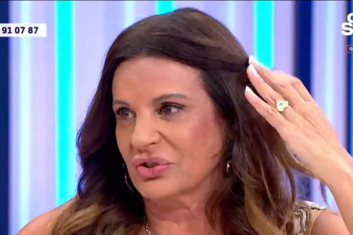 Dona de cabell castany llarg parlant en un programa de televisió amb un fons de llums blaves i blanques i mostrant un anell a la mà dreta