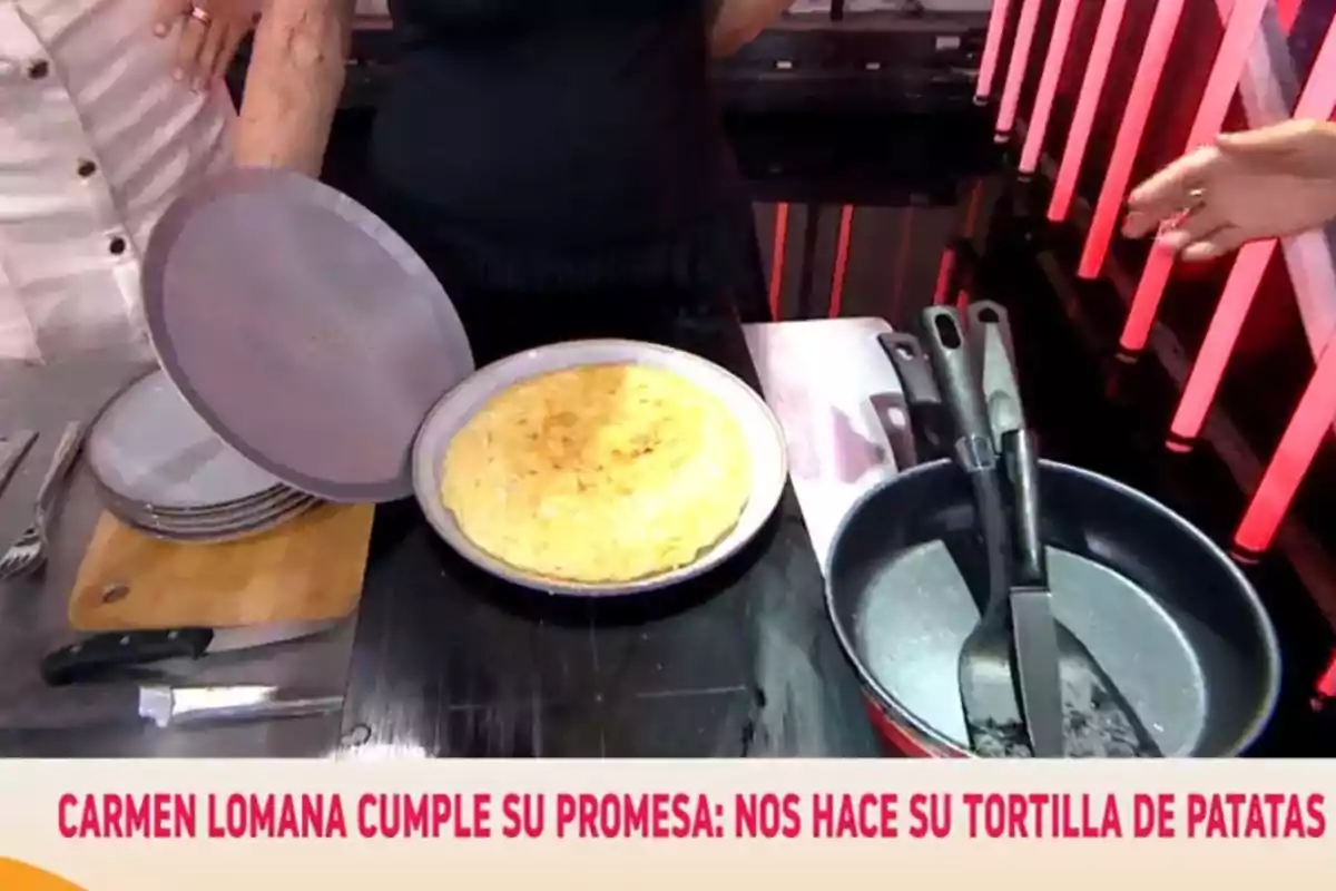 Una tortilla de patatas recién hecha está servida en un plato sobre una mesa junto a una sartén, utensilios de cocina y varios platos apilados Una tortilla de patatas recién hecha está servida en un plato sobre una mesa junto a una sartén, utensilios de cocina y varios platos apilados