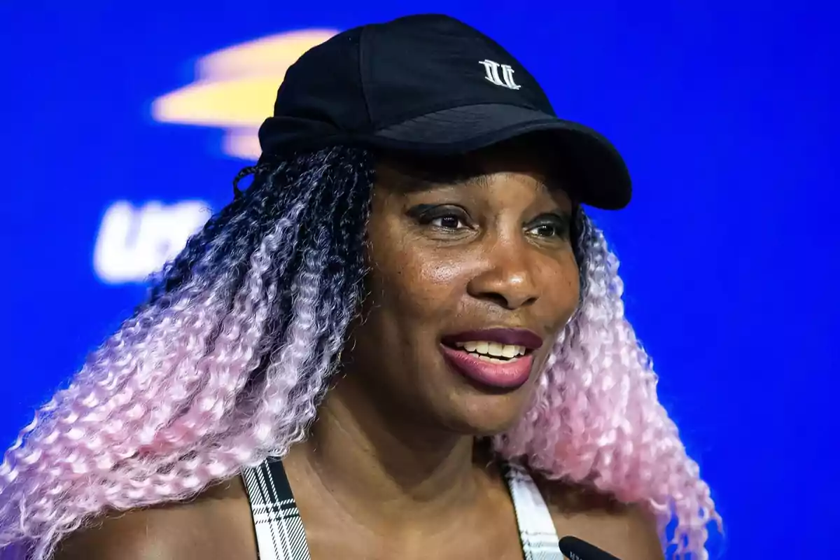 Venus Williams con gorra negra y cabello rizado en tonos oscuros y lilas sonriendo frente a un fondo azul Venus Williams con gorra negra y cabello rizado en tonos oscuros y lilas sonriendo frente a un fondo azul