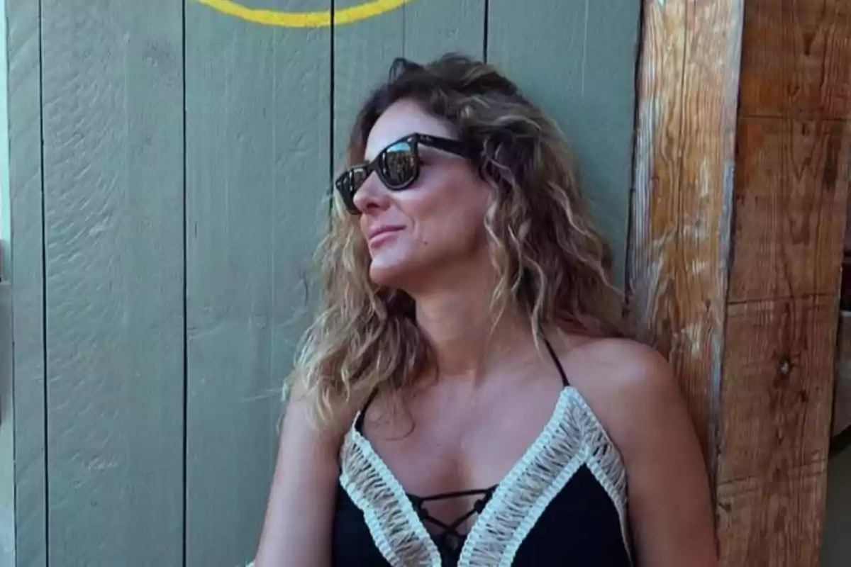 Verónica Dulanto con gafas de sol y cabello rizado sentada junto a una pared de madera, usando un vestido negro con detalles claros y mirando hacia un lado Verónica Dulanto con gafas de sol y cabello rizado sentada junto a una pared de madera, usando un vestido negro con detalles claros y mirando hacia un lado