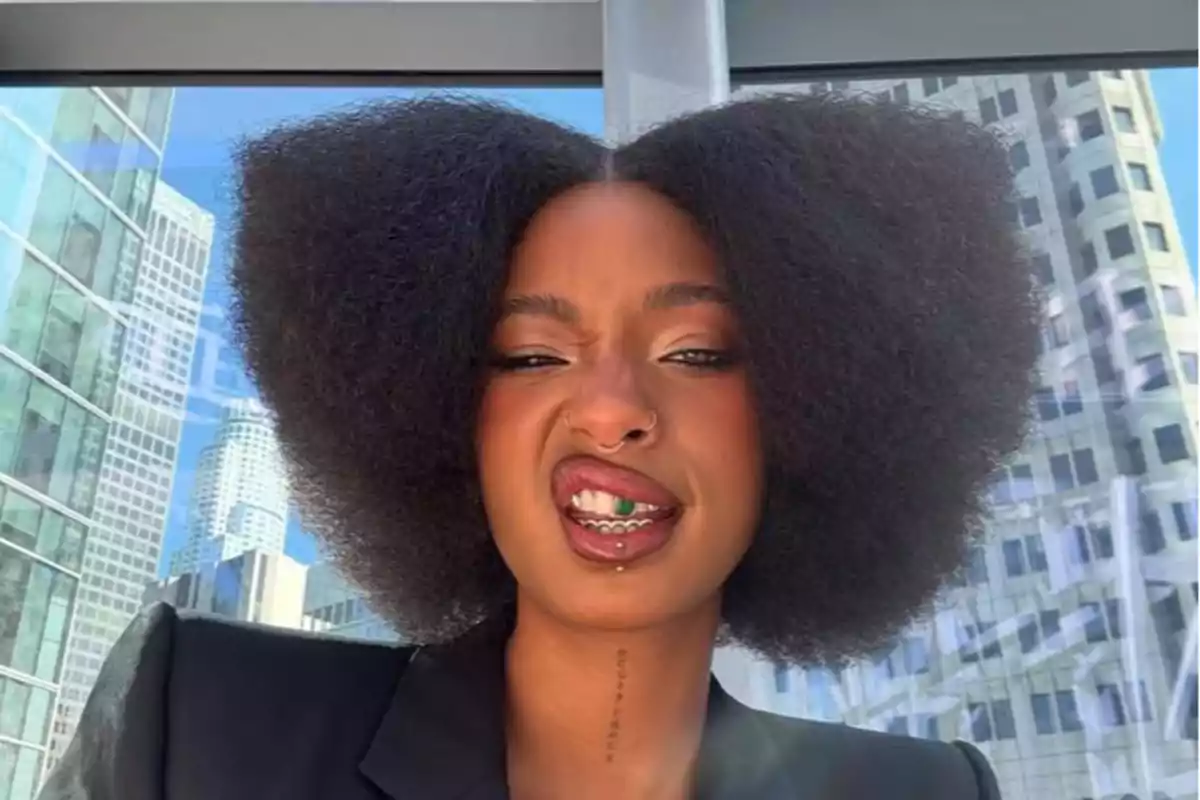 Persona amb cabell afro voluminós en forma de cor fent una ganyota divertida davant d’una finestra amb edificis de fons Persona amb cabell afro voluminós en forma de cor fent una ganyota divertida davant d’una finestra amb edificis de fons