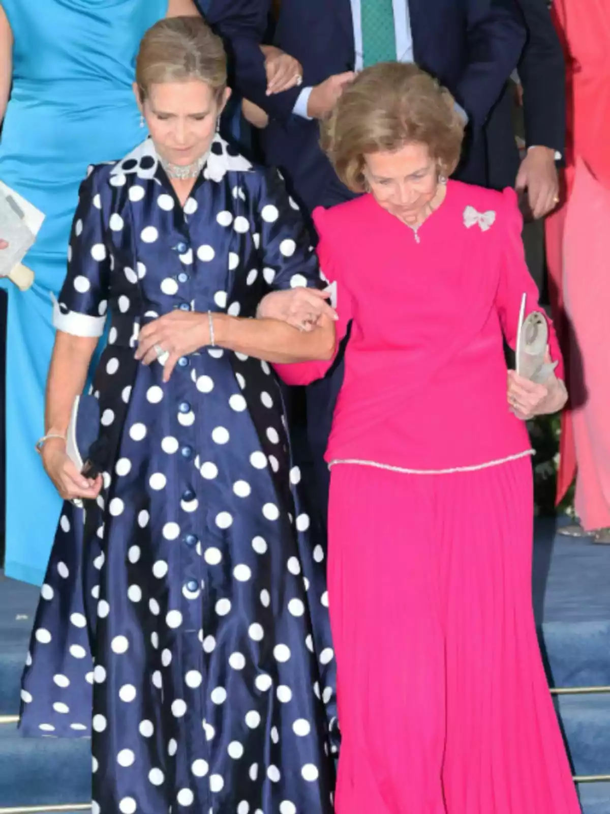La infanta Elena i la reina Sofia, elegantment vestides, baixen unes escales agafades del braç; una porta un vestit blau marí amb topos blancs i l’altra un conjunt fúcsia. La infanta Elena i la reina Sofia, elegantment vestides, baixen unes escales agafades del braç; una porta un vestit blau marí amb topos blancs i l’altra un conjunt fúcsia.