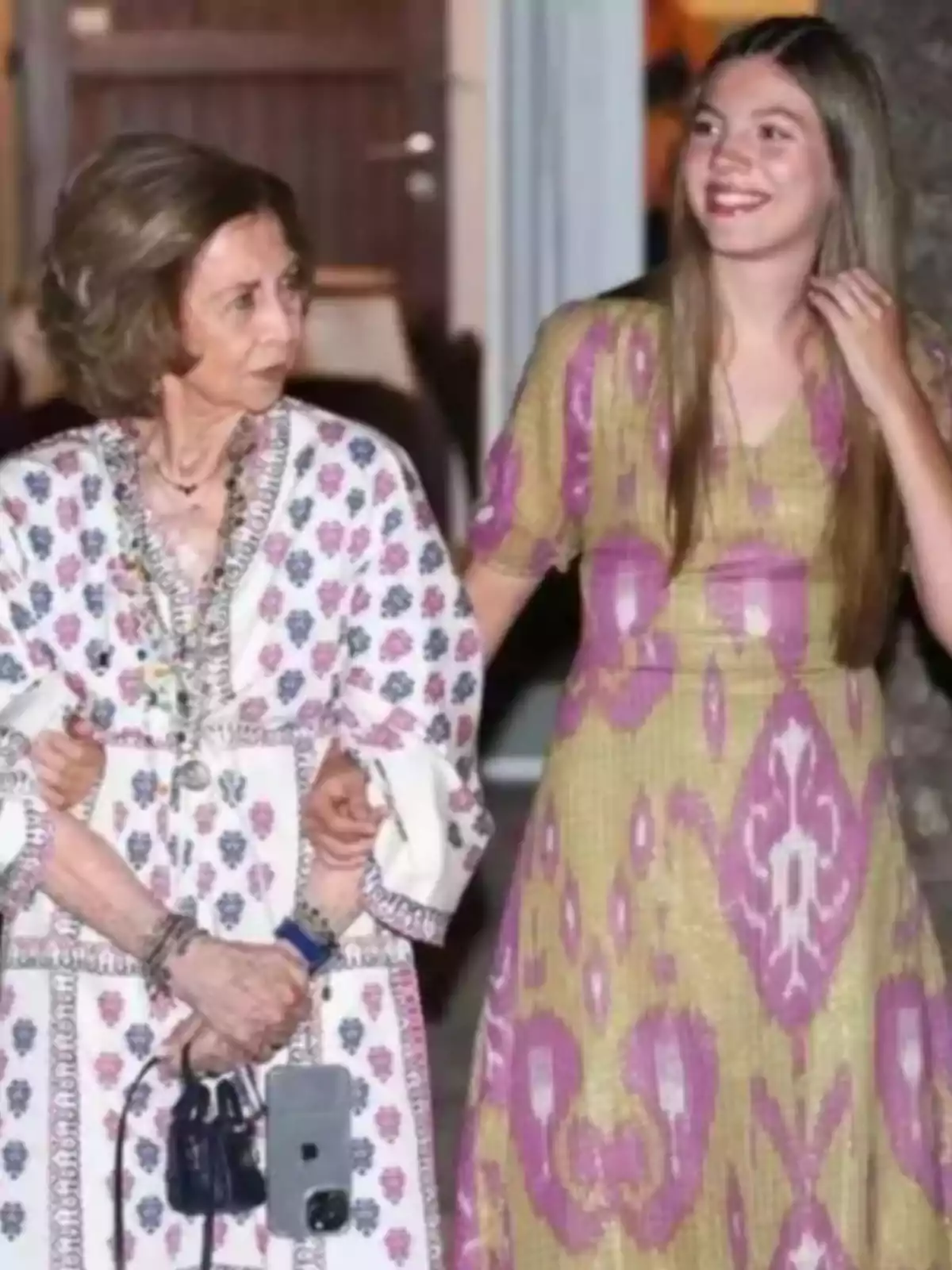 Dos mujeres caminan tomadas del brazo, una de ellas mayor con vestido blanco estampado y la otra joven con vestido largo amarillo y rosa, ambas sonríen y parecen disfrutar el momento juntas Dos mujeres caminan tomadas del brazo, una de ellas mayor con vestido blanco estampado y la otra joven con vestido largo amarillo y rosa, ambas sonríen y parecen disfrutar el momento juntas