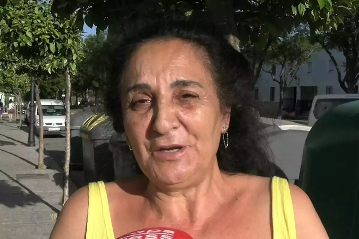 Mujer de cabello oscuro y rizado, que es Inma -la madre de Michu-, con vestido amarillo hablando frente a un micrófono rojo en una calle arbolada y soleada. Mujer de cabello oscuro y rizado, que es Inma -la madre de Michu-, con vestido amarillo hablando frente a un micrófono rojo en una calle arbolada y soleada.