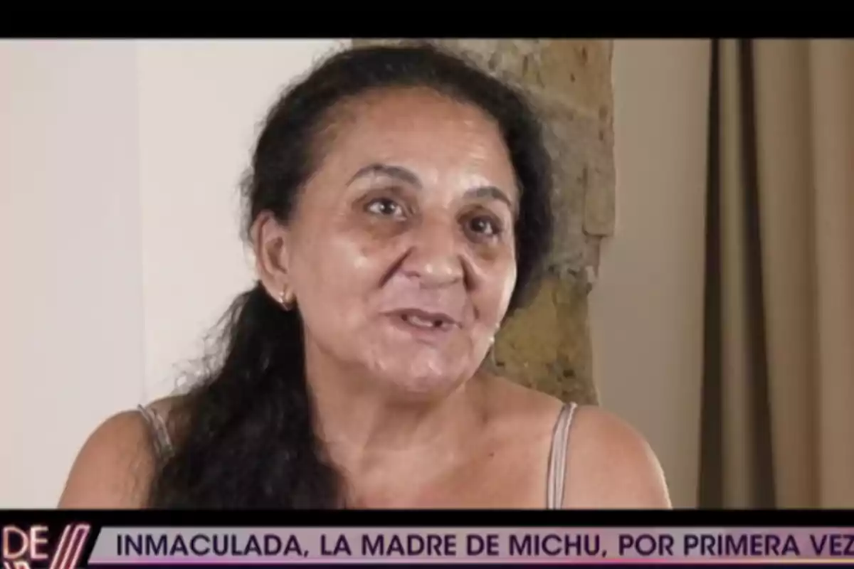 Inmaculada, con expresión sonriente, sentada frente a una pared clara y una cortina, con un rótulo en la parte inferior que menciona a Inmaculada, la madre de Michu. Inmaculada, con expresión sonriente, sentada frente a una pared clara y una cortina, con un rótulo en la parte inferior que menciona a Inmaculada, la madre de Michu.