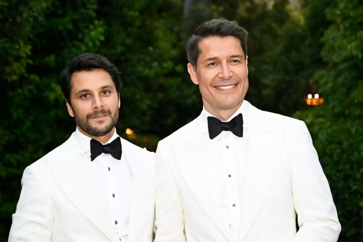 Miguel García y Jaime Cantizano vestidos con esmoquin blanco y pajarita negra posan juntos al aire libre con árboles de fondo Miguel García y Jaime Cantizano vestidos con esmoquin blanco y pajarita negra posan juntos al aire libre con árboles de fondo