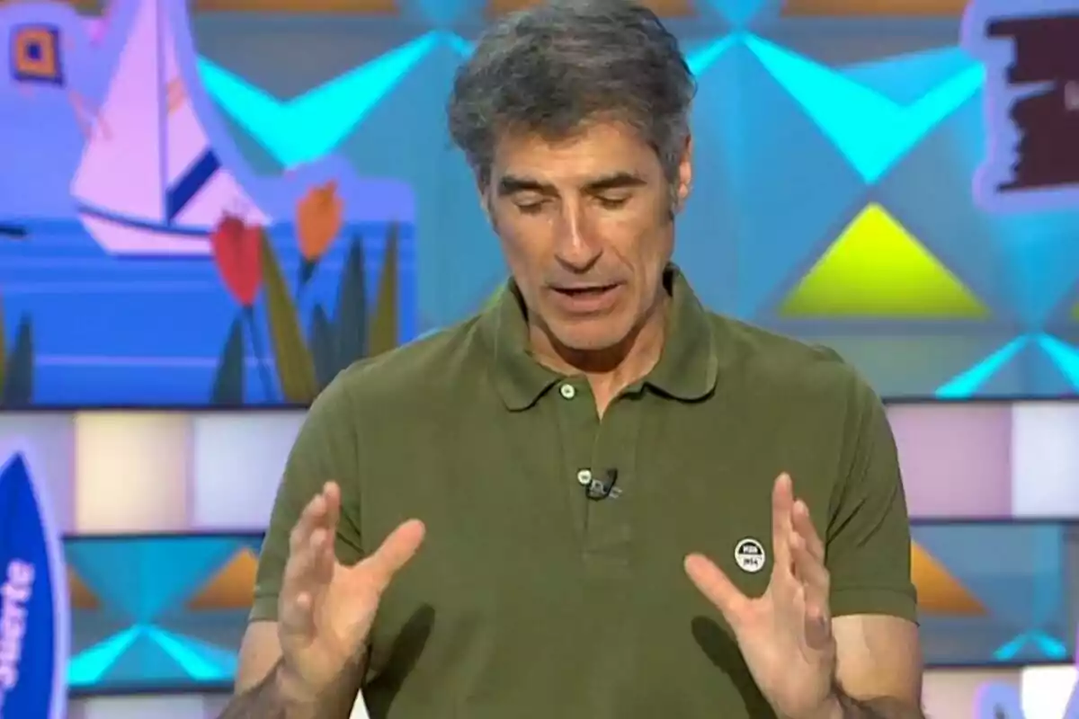 Jorge Fernández vestido con camisa polo verde hablando y gesticulando con las manos en un set colorido con fondo de figuras geométricas y dibujos de flores y un barco. Jorge Fernández vestido con camisa polo verde hablando y gesticulando con las manos en un set colorido con fondo de figuras geométricas y dibujos de flores y un barco.
