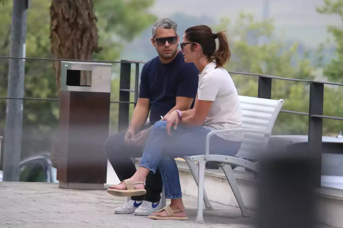 José Fernando i una dona asseguts en un banc a l’aire lliure conversen mentre porten ulleres de sol envoltats d’arbres i una paperera propera.