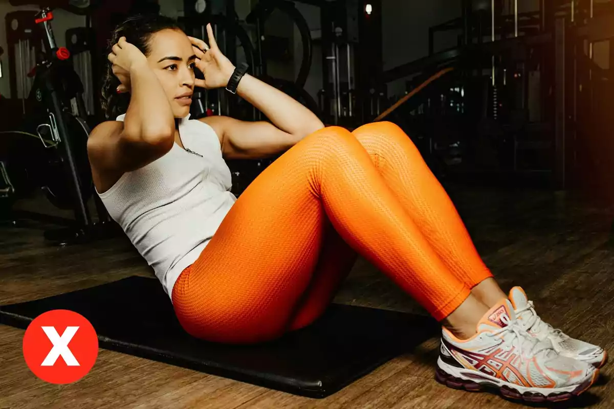 Mujer haciendo abdominales en un gimnasio con ropa deportiva naranja y un símbolo de equis roja en la esquina inferior izquierda Mujer haciendo abdominales en un gimnasio con ropa deportiva naranja y un símbolo de equis roja en la esquina inferior izquierda