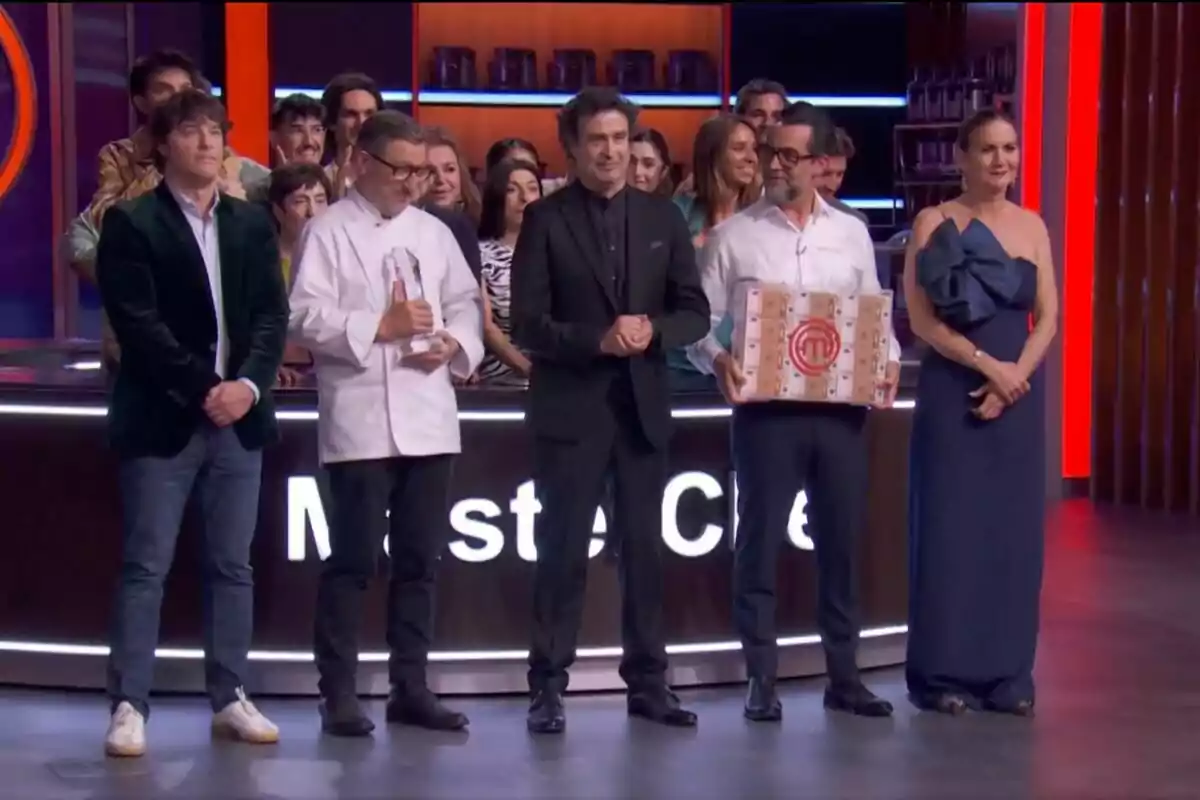 Grup de persones —que són el jurat de MasterChef— en un plató de televisió d’un concurs de cuina, alguns sostenen un trofeu i una caixa amb el logotip del programa, mentre la resta aplaudeix i somriu al fons. Grup de persones —que són el jurat de MasterChef— en un plató de televisió d’un concurs de cuina, alguns sostenen un trofeu i una caixa amb el logotip del programa, mentre la resta aplaudeix i somriu al fons.