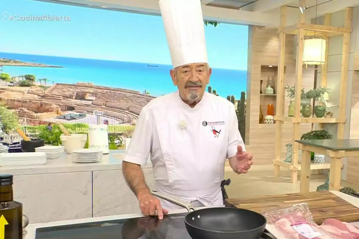 Xef amb barret alt de cuina en un plató de programa culinari amb vista al mar i ruïnes al fons, que és Karlos Arguiñano, envoltat d’estris i plats, al costat d’una paella i carn sobre la taula. Xef amb barret alt de cuina en un plató de programa culinari amb vista al mar i ruïnes al fons, que és Karlos Arguiñano, envoltat d’estris i plats, al costat d’una paella i carn sobre la taula.