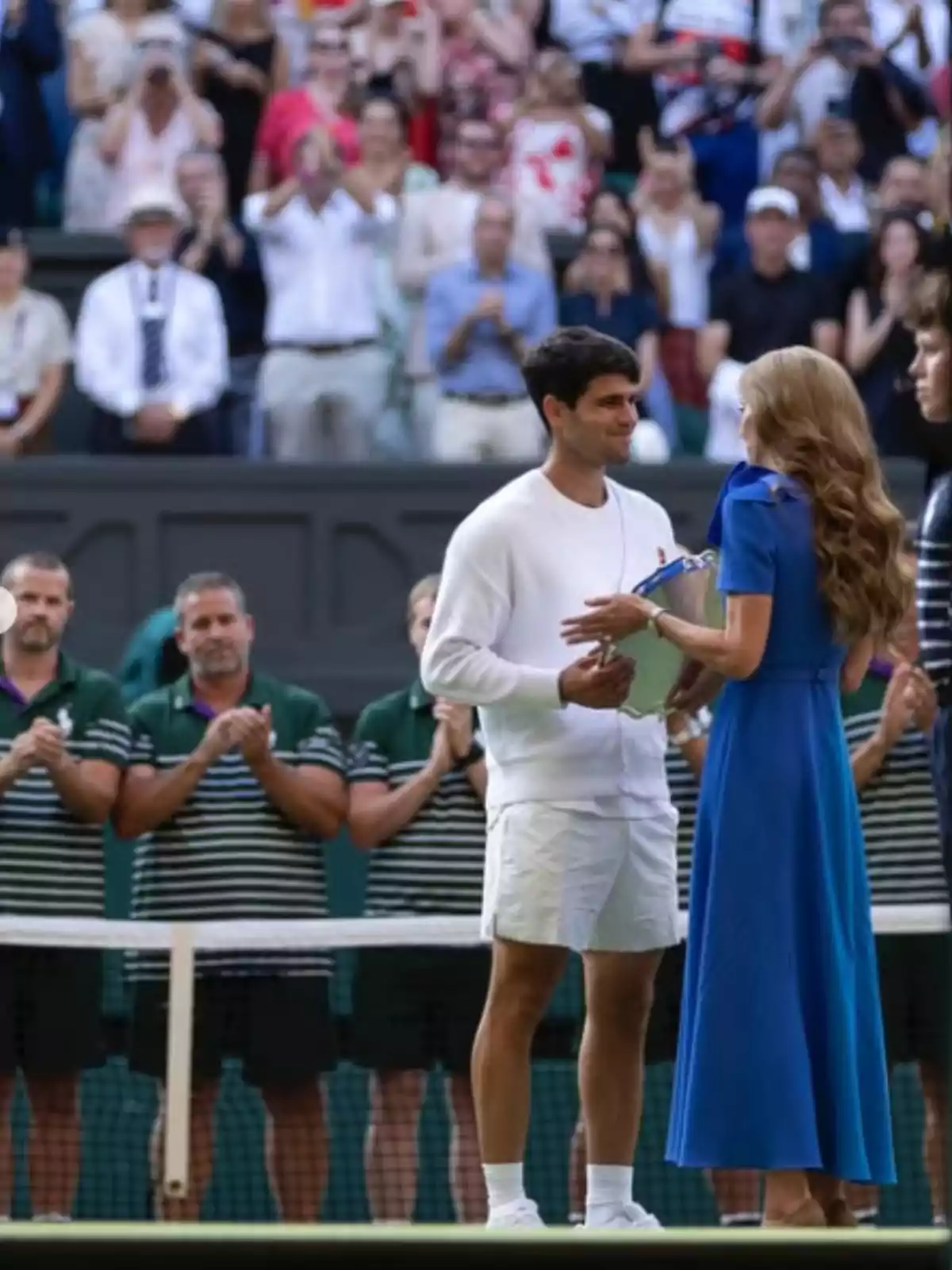 Un tennista rep un trofeu d’una dona vestida de blau en una pista de tennis mentre el públic i l’equip aplaudeixen al fons. Un tennista rep un trofeu d’una dona vestida de blau en una pista de tennis mentre el públic i l’equip aplaudeixen al fons.