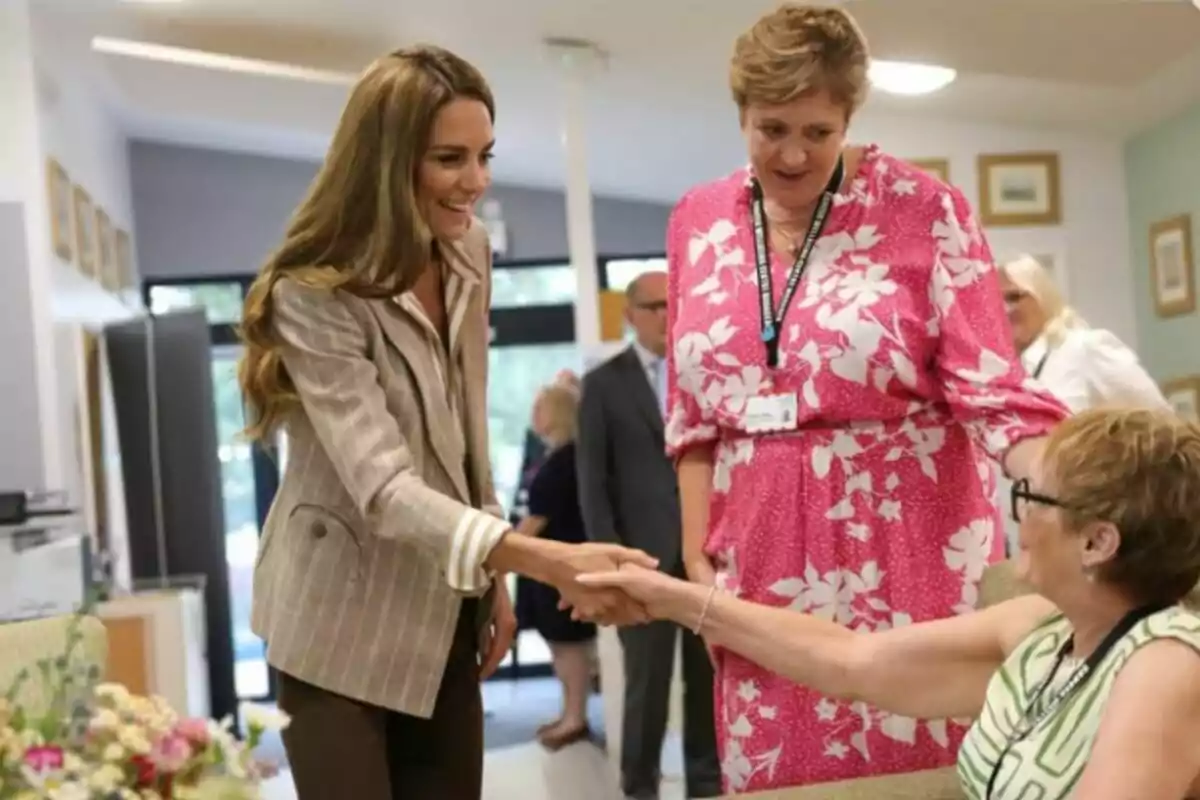 Una dona de cabells llargs i solts saluda amb la mà una senyora asseguda mentre una altra dona amb vestit rosa les observa somrient en una sala il·luminada. Una dona de cabells llargs i solts saluda amb la mà una senyora asseguda mentre una altra dona amb vestit rosa les observa somrient en una sala il·luminada.