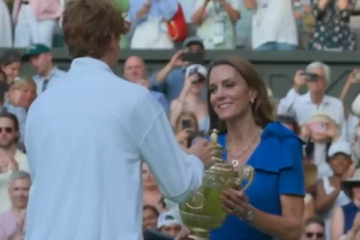 A woman in a blue dress, who is Kate Middleton, presents a golden trophy to a tennis player while the crowd watches and takes photos at Wimbledon. A woman in a blue dress, who is Kate Middleton, presents a golden trophy to a tennis player while the crowd watches and takes photos at Wimbledon.