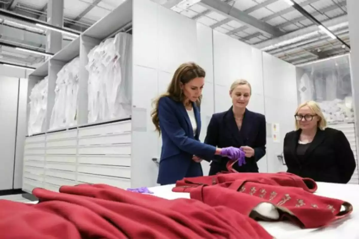 Tres dones observen i manipulen acuradament peces de roba vermelles antigues sobre una taula en una sala d’emmagatzematge moderna i ben il·luminada.