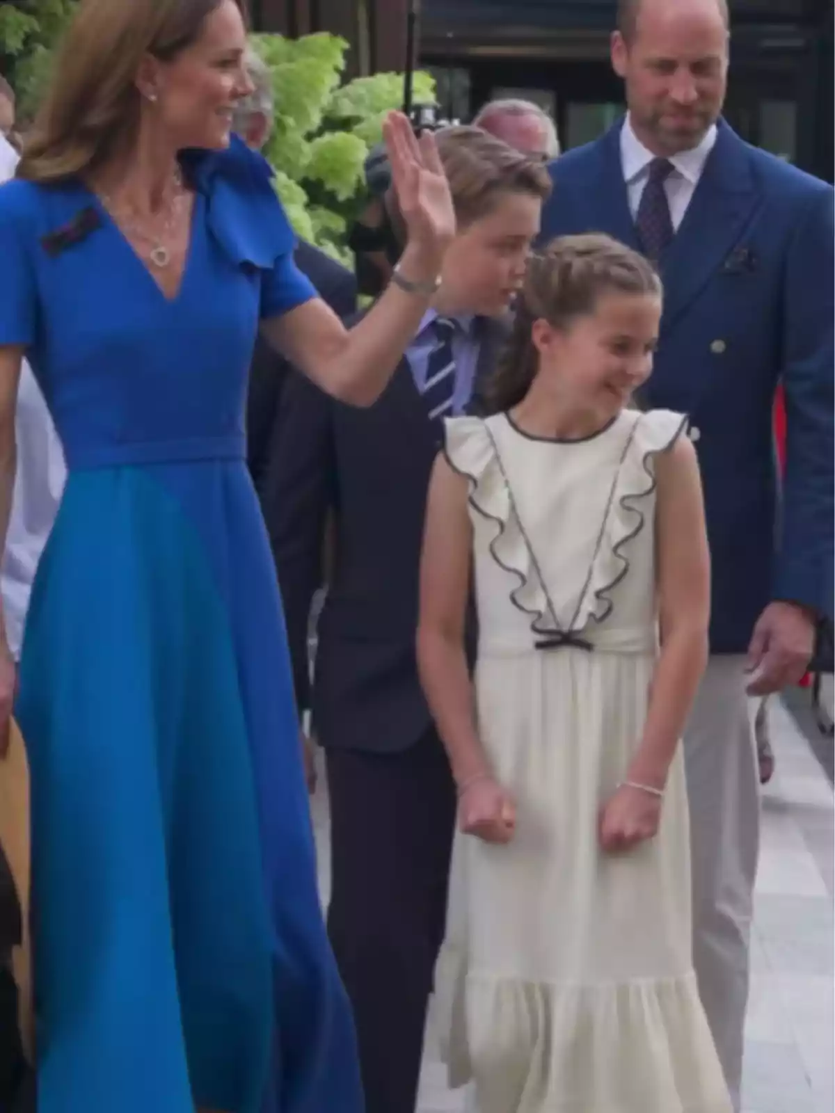 Una dona amb vestit blau saluda mentre està acompanyada d’un home amb americana blava, un nen amb vestit i una nena amb vestit blanc, tots somriuen i semblen estar en un esdeveniment a l’aire lliure. Una dona amb vestit blau saluda mentre està acompanyada d’un home amb americana blava, un nen amb vestit i una nena amb vestit blanc, tots somriuen i semblen estar en un esdeveniment a l’aire lliure.