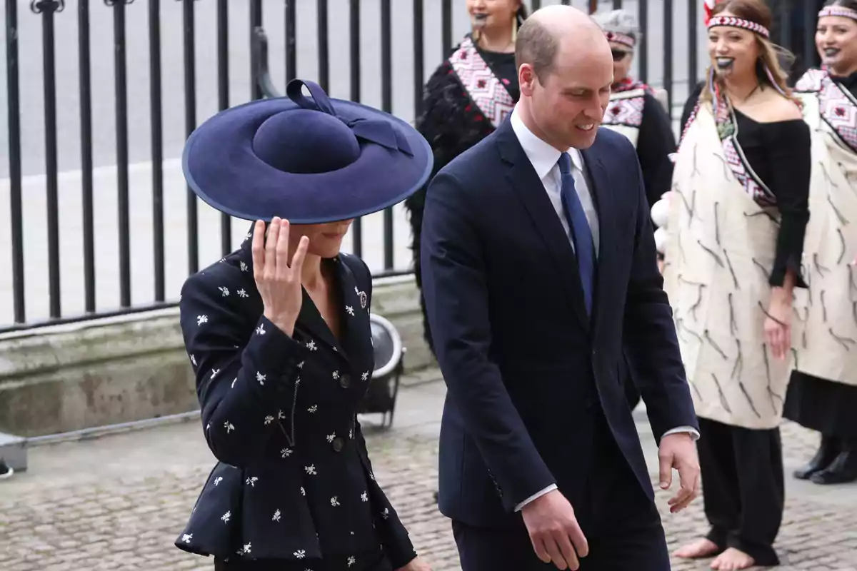 Kate Middleton amb un gran barret blau camina al costat del príncep Guillem amb vestit fosc mentre diverses persones amb vestimenta tradicional somriuen al fons. Kate Middleton amb un gran barret blau camina al costat del príncep Guillem amb vestit fosc mentre diverses persones amb vestimenta tradicional somriuen al fons.