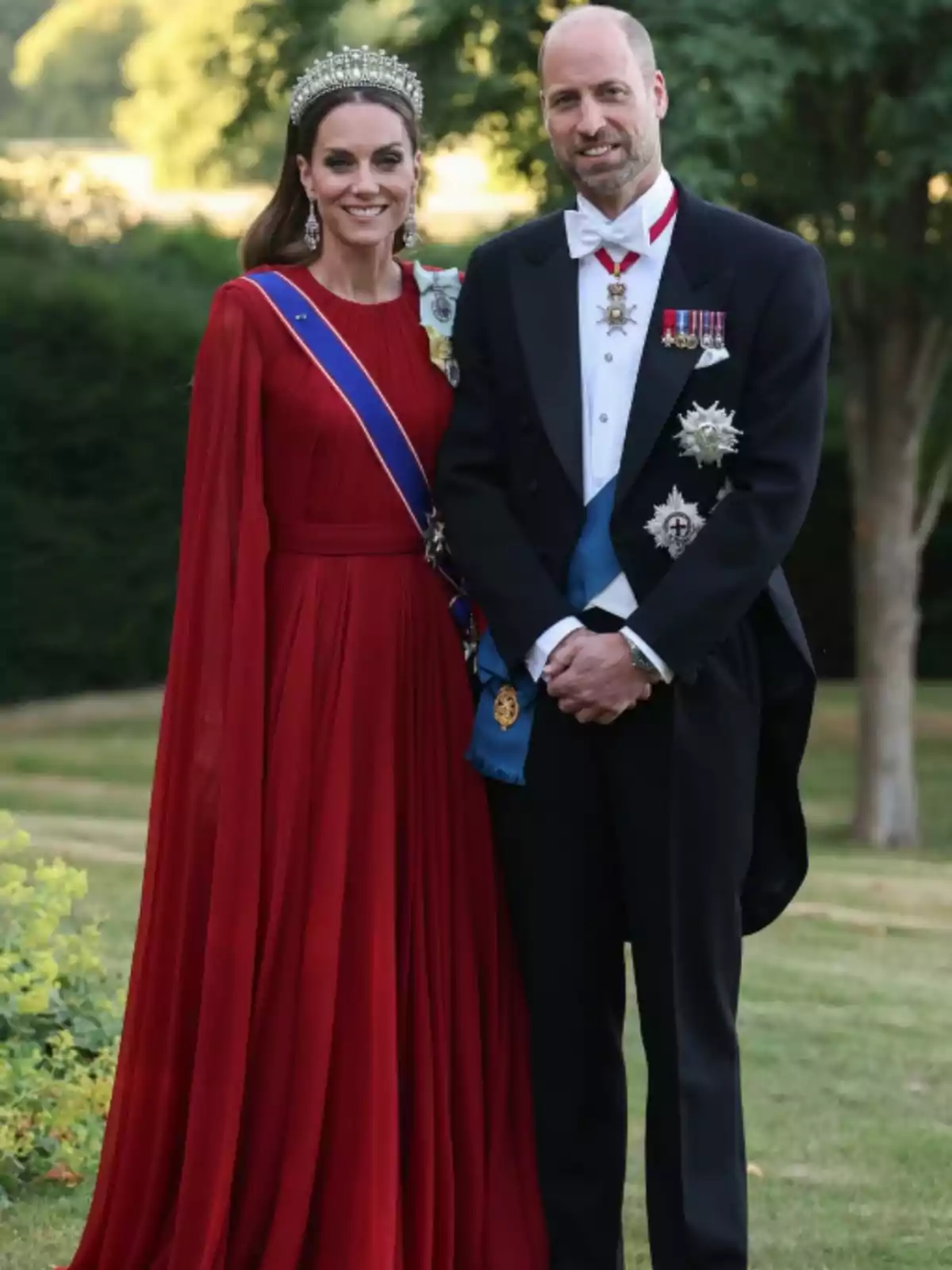 Una dona amb vestit vermell llarg i tiara al costat d’un home amb esmòquing i condecoracions posen a l’aire lliure en un jardí. Una dona amb vestit vermell llarg i tiara al costat d’un home amb esmòquing i condecoracions posen a l’aire lliure en un jardí.