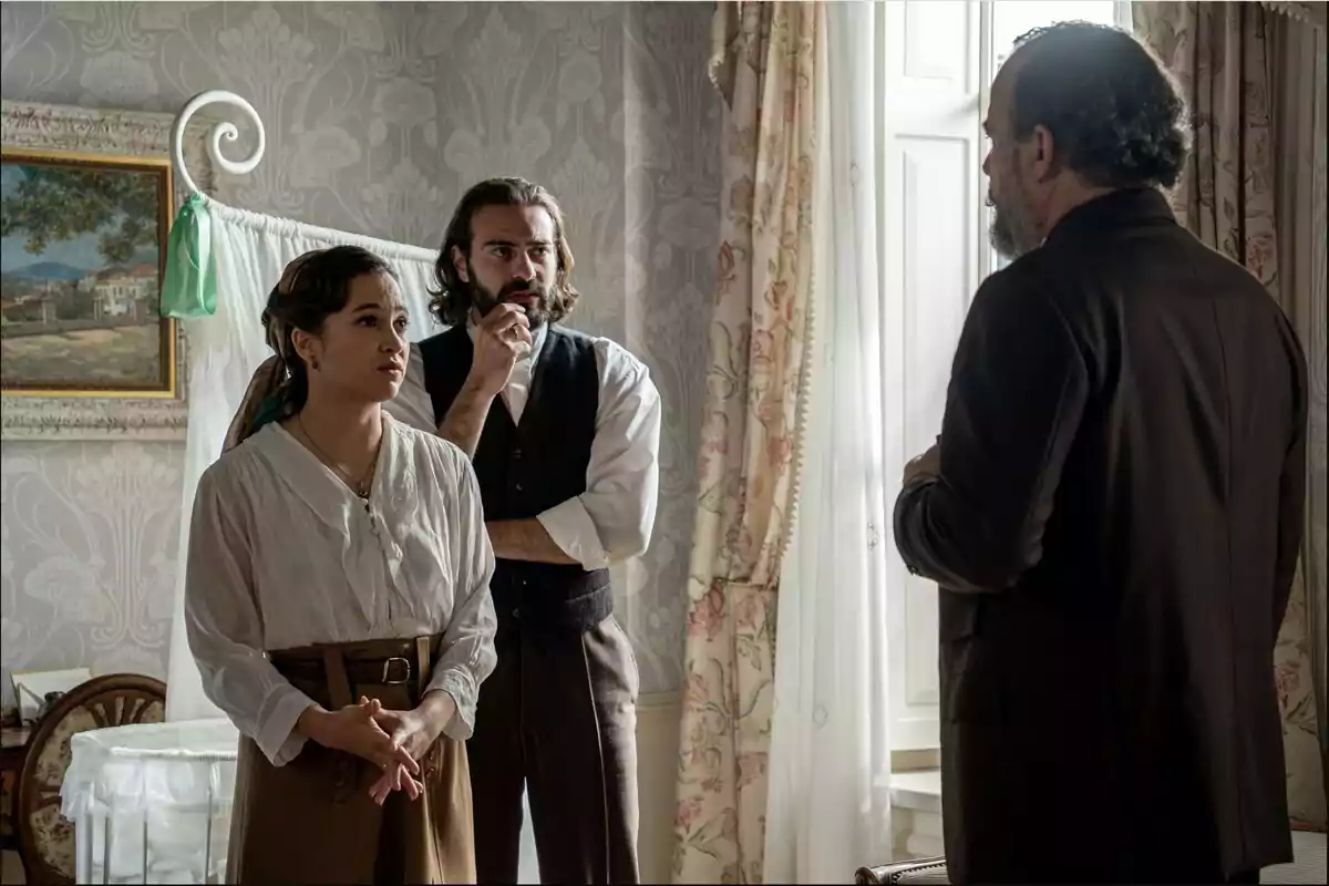Catalina, Adriano i Alonso conversen en una habitació elegant amb cortines i paper pintat, dues d’elles miren atentament un home d’esquena al costat d’una finestra il·luminada. Captura de ‘La Promesa’ Catalina, Adriano i Alonso conversen en una habitació elegant amb cortines i paper pintat, dues d’elles miren atentament un home d’esquena al costat d’una finestra il·luminada. Captura de ‘La Promesa’