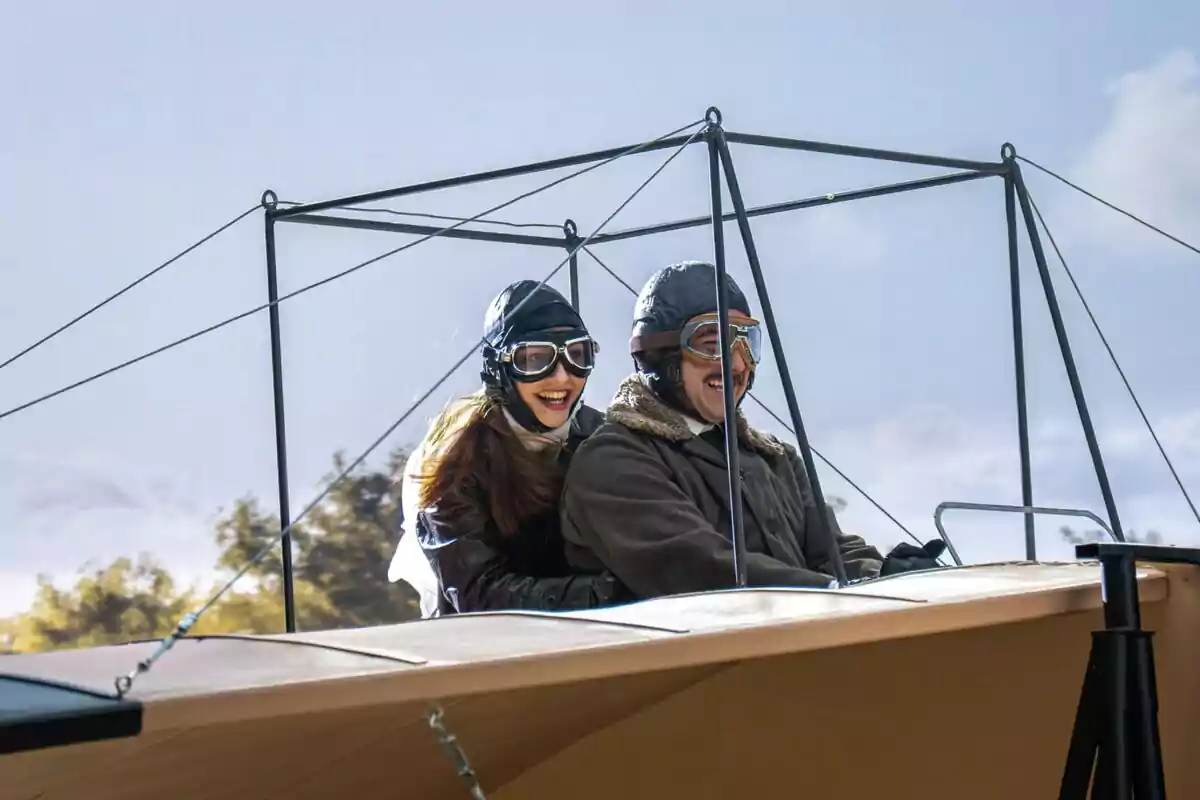 Enora i Manuel somrients amb ulleres i gorres d’aviador gaudeixen d’un vol en un avió antic sota un cel serè. Captura de ‘La Promesa’ Enora i Manuel somrients amb ulleres i gorres d’aviador gaudeixen d’un vol en un avió antic sota un cel serè. Captura de ‘La Promesa’