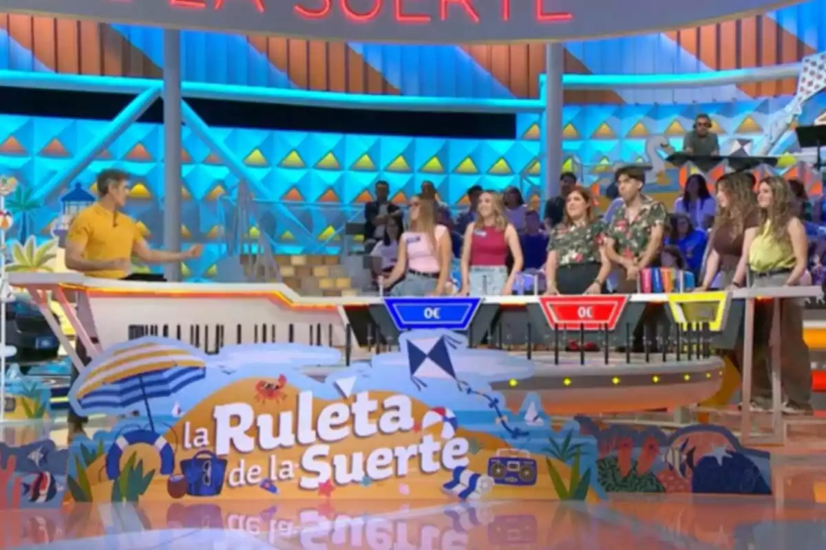 Un grup de concursants i Jorge Fernández participen al plató acolorit del programa de televisió La Ruleta de la Suerte amb decoració d’estiu i públic al fons. Captura de 'La Ruleta de la Suerte' Un grup de concursants i Jorge Fernández participen al plató acolorit del programa de televisió La Ruleta de la Suerte amb decoració d’estiu i públic al fons. Captura de 'La Ruleta de la Suerte'