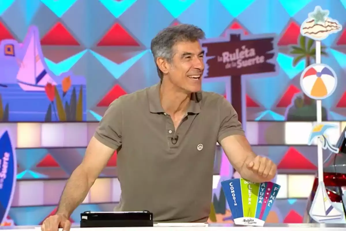 Hombre sonriente de cabello canoso con camiseta polo beige en un set colorido de programa de televisión con letrero de La Ruleta de la Suerte al fondo Hombre sonriente de cabello canoso con camiseta polo beige en un set colorido de programa de televisión con letrero de La Ruleta de la Suerte al fondo