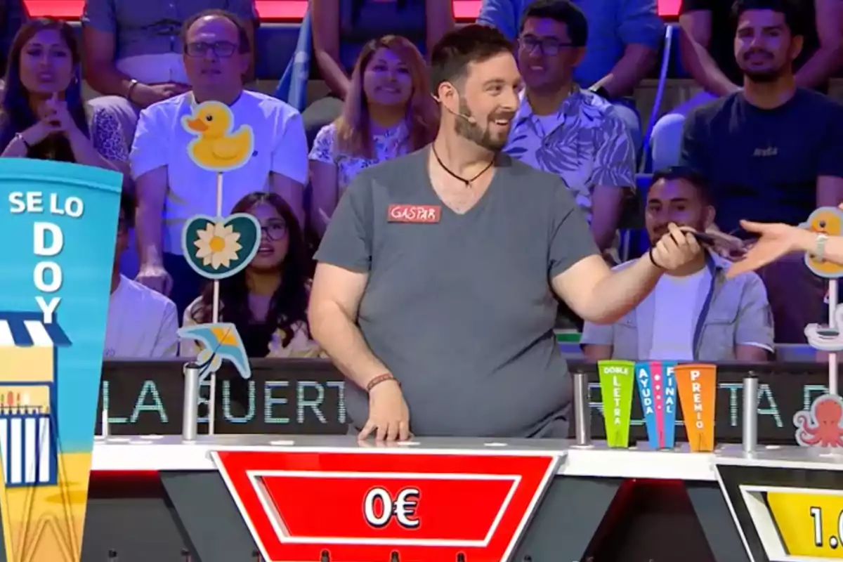 Un concursante sonriente en un programa de televisión recibe un objeto de otra persona mientras el público observa y el marcador muestra cero euros Un concursante sonriente en un programa de televisión recibe un objeto de otra persona mientras el público observa y el marcador muestra cero euros