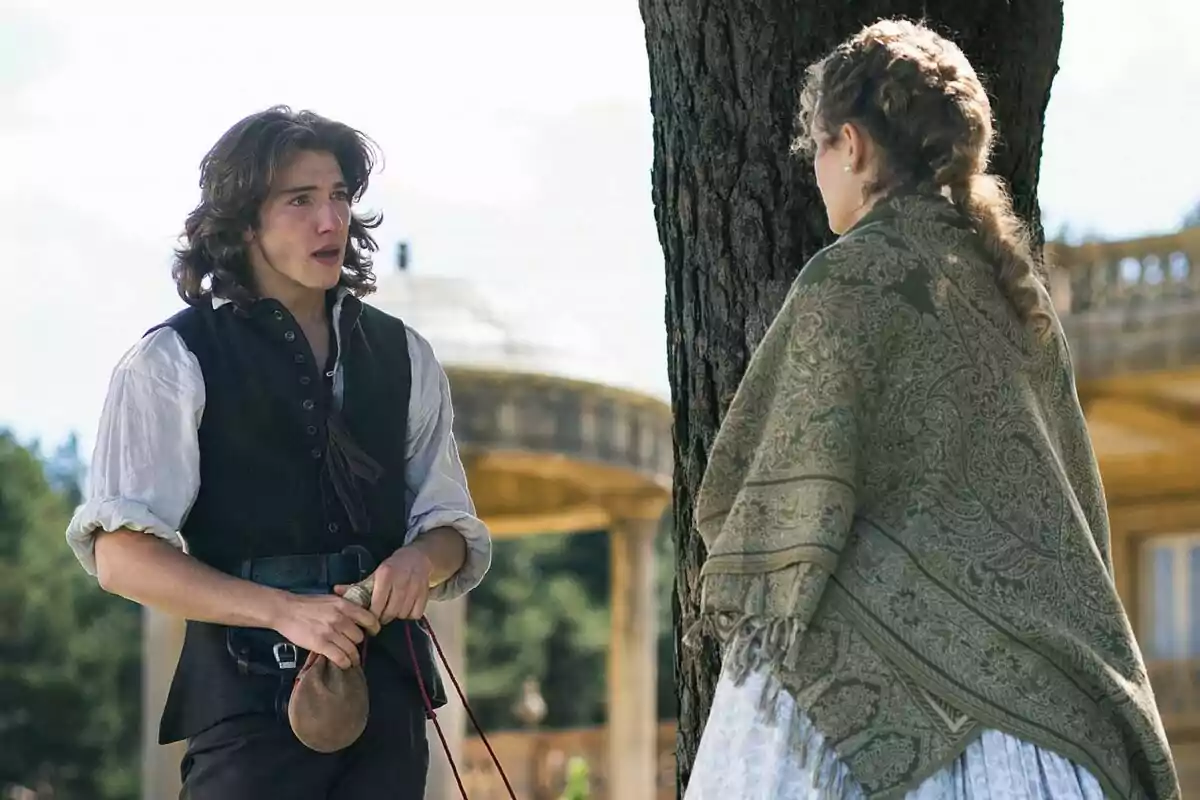 Julen Katzy con cabello largo y Emma Guilera con trenza conversan junto a un árbol en un entorno al aire libre con vestimenta de época en Valle Salvaje Julen Katzy con cabello largo y Emma Guilera con trenza conversan junto a un árbol en un entorno al aire libre con vestimenta de época en Valle Salvaje