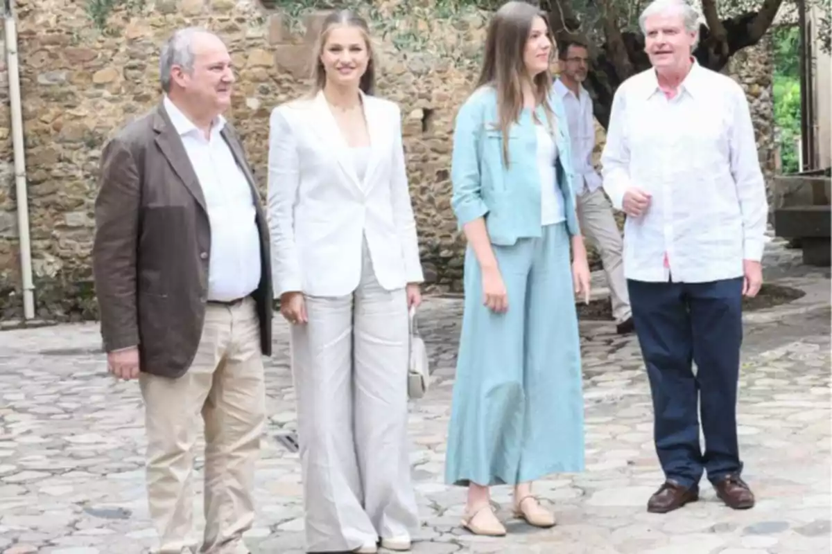 Quatre persones posen dretes sobre un camí de pedra davant d’una paret de pedra, dos homes grans als extrems i la princesa Leonor i la infanta Sofia al centre, tots vestits amb roba formal i colors clars.