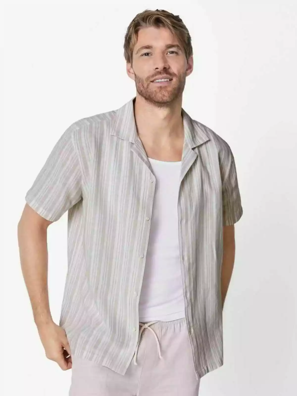 Home jove amb barba i cabells rossos porta una camisa de màniga curta de ratlles sobre una samarreta blanca i pantalons clars posant davant d’un fons blanc Home jove amb barba i cabells rossos porta una camisa de màniga curta de ratlles sobre una samarreta blanca i pantalons clars posant davant d’un fons blanc