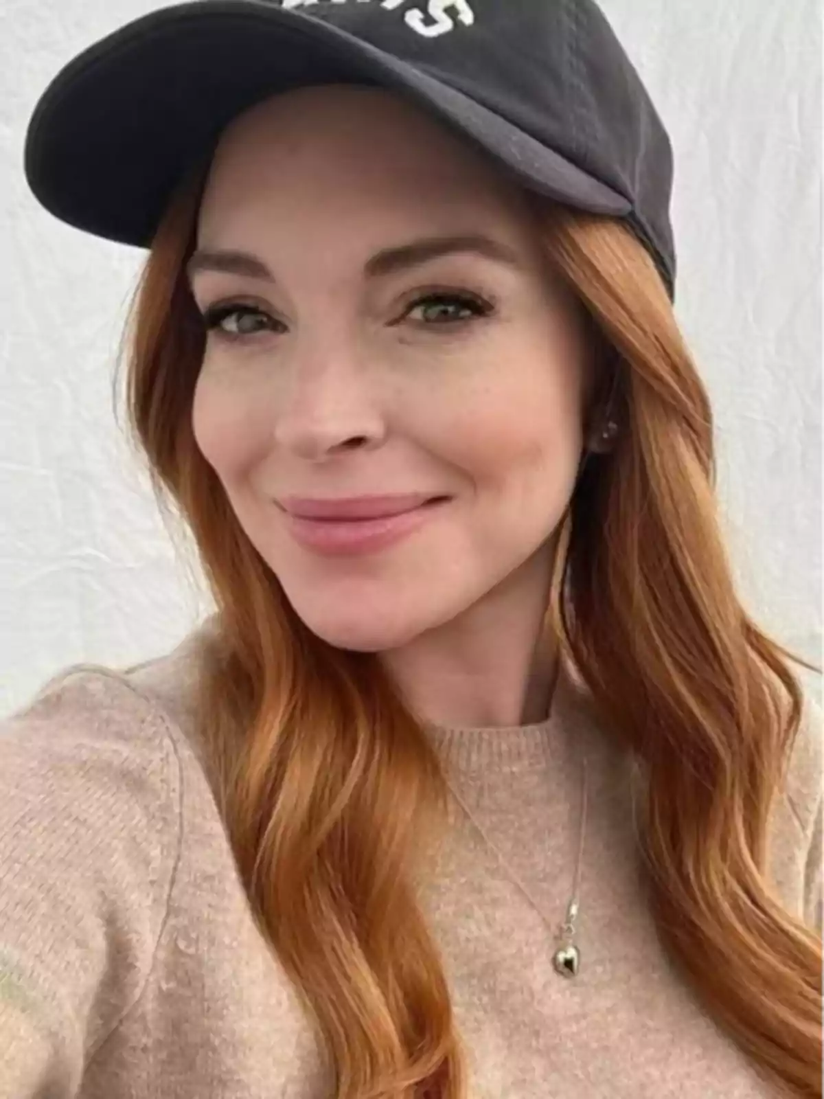 Lindsay Lohan wearing a beige sweater and a black cap smiling in front of a light background Lindsay Lohan wearing a beige sweater and a black cap smiling in front of a light background
