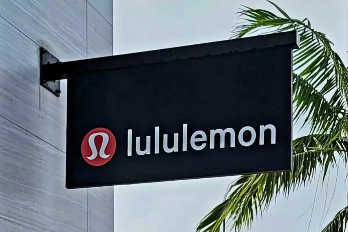 Letrero negro de la tienda lululemon con el logo rojo y letras blancas junto a una palmera Letrero negro de la tienda lululemon con el logo rojo y letras blancas junto a una palmera