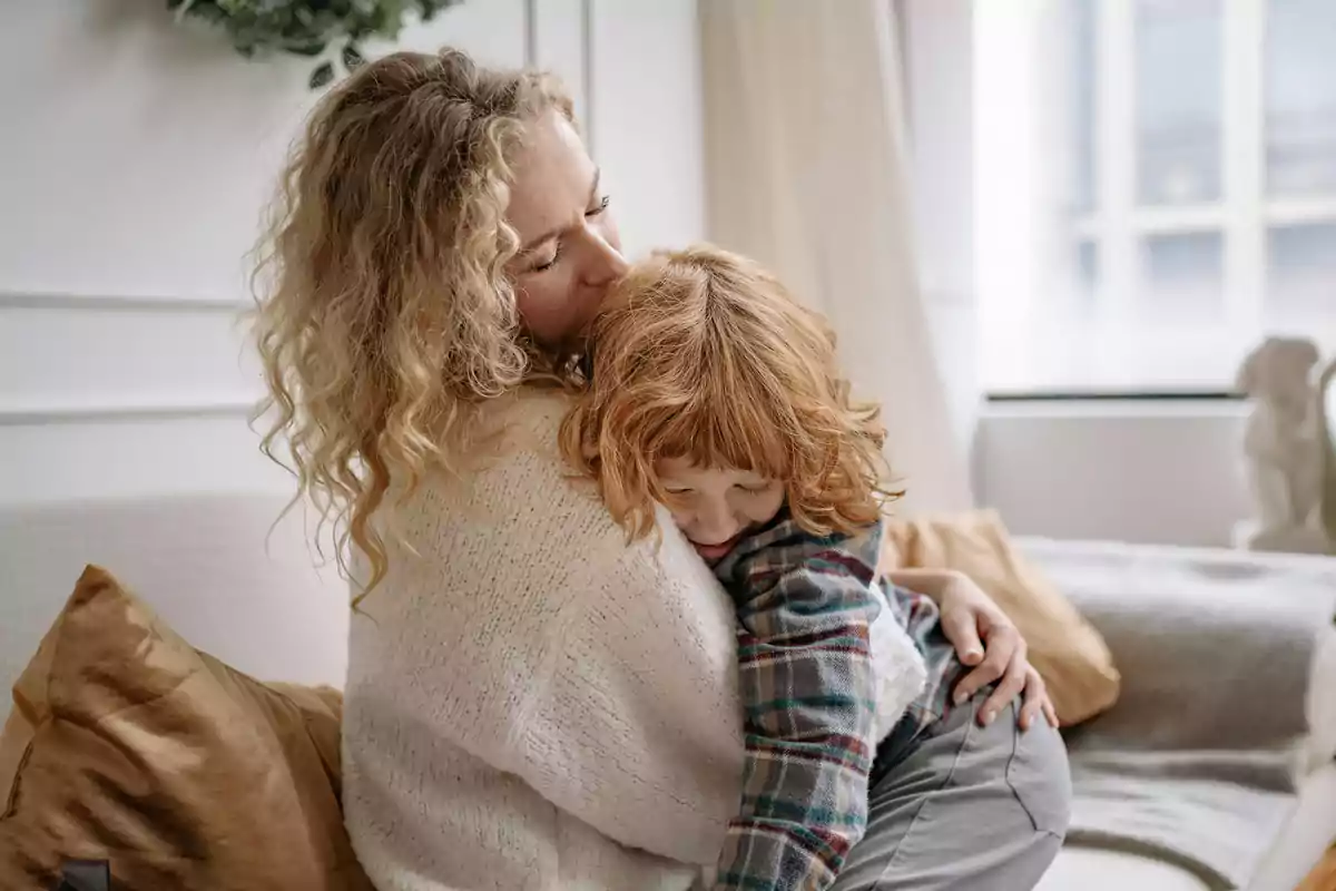 Dona abraçant i fent un petó a un nen petit en un sofà en una sala il·luminada Dona abraçant i fent un petó a un nen petit en un sofà en una sala il·luminada