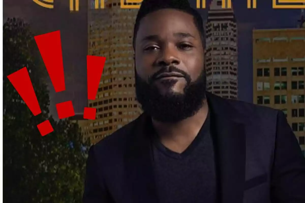 Man with a beard and short hair dressed in black in front of buildings with red exclamation marks next to him