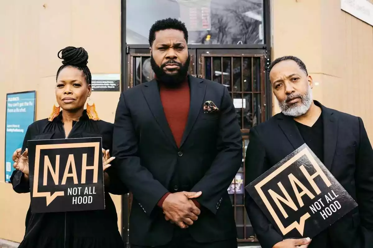 Three people pose in front of a building, two of them holding signs that say NAH NOT ALL HOOD, and all of them are wearing dark formal clothing. Three people pose in front of a building, two of them holding signs that say NAH NOT ALL HOOD, and all of them are wearing dark formal clothing.