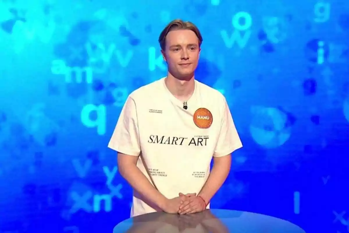 Manu Pascual, dret darrere d’un faristol en un plató de televisió amb fons blau i lletres disperses, porta una samarreta blanca amb el text SMART ART i un cercle taronja que diu MANU. Manu Pascual, dret darrere d’un faristol en un plató de televisió amb fons blau i lletres disperses, porta una samarreta blanca amb el text SMART ART i un cercle taronja que diu MANU.