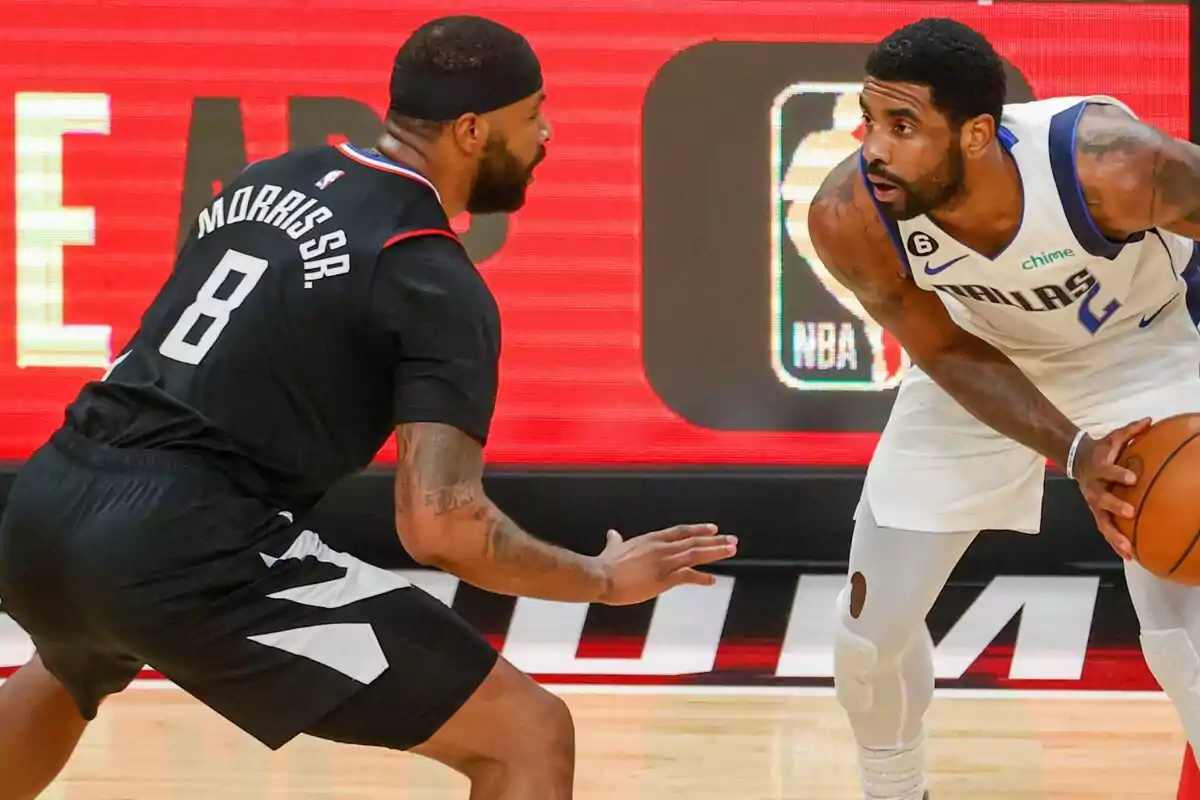 Dos jugadors de bàsquet competeixen durant un partit, un defensant i l’altre sostenint la pilota mentre es miren fixament. Dos jugadors de bàsquet competeixen durant un partit, un defensant i l’altre sostenint la pilota mentre es miren fixament.