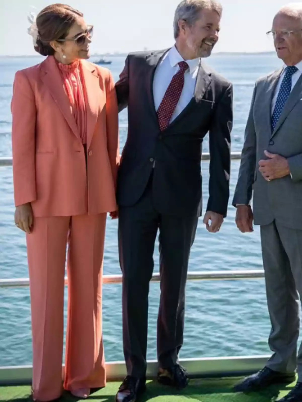 Tres persones elegantment vestides conversen i somriuen dretes sobre una plataforma vora el mar en un dia assolellat. Tres persones elegantment vestides conversen i somriuen dretes sobre una plataforma vora el mar en un dia assolellat.